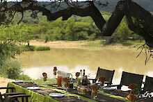 Ant´s Africa Safaris Flußlandschaft, gedeckter Tisch unter Baum mit bewaldeten Ufer im Hintergrund