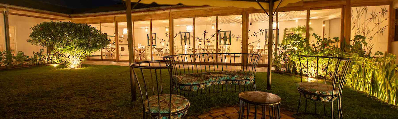 Tansania Mama Iringa Garten mit eleganter Sitzgruppe und Blick auf beleuchteten Restaurant mit Glasfront