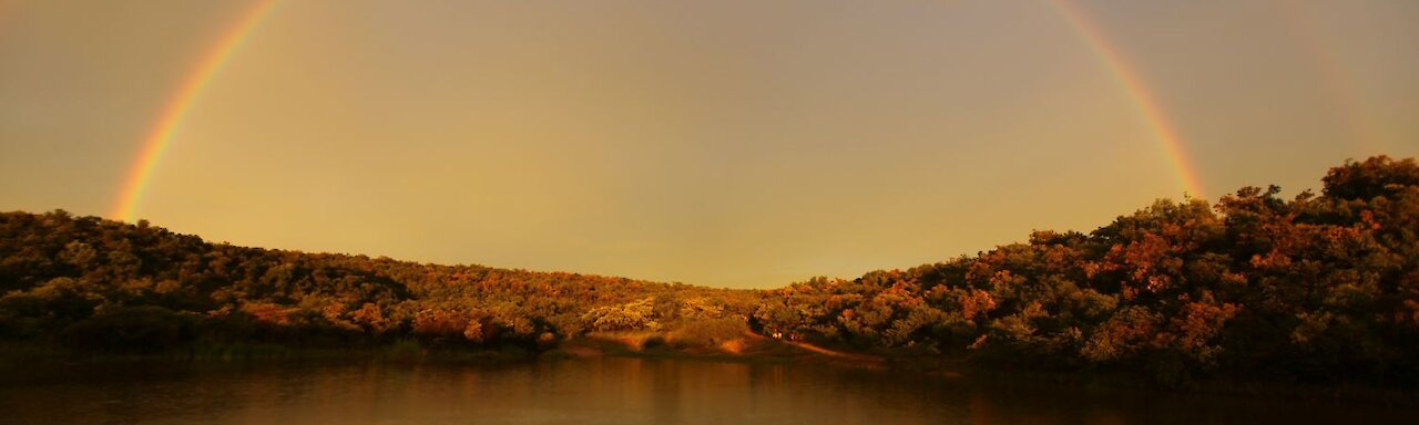 Ant´s Africa Safaris - Regenbogen über Flusslandschaft mit Reiter und Pferden am bewaldeten Ufer im Hintergrund. Südafrika